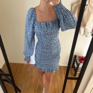 Blommig blå kortklänning med volang - Supersöt blå kortklänning med vitt blommönster och volangkant längst ner. Klänningen har långärmade puffärmar med smock vid handlederna och en fyrkantig urringning både fram och bak. Väldigt stretchig i materialet och har även en dragkedja i sidan som gör den lätt att ha på och av. 