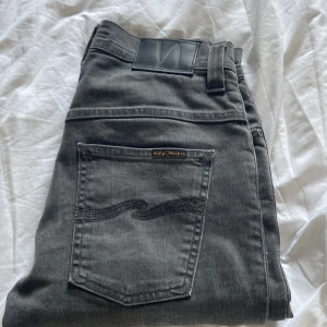 Grå jeans från Nudie Jeans, W27 L32 - Snygga grå jeans från Nudie Jeans med klassisk bakficka och broderad vågdetalj. Jeansen har en normal passform och är tillverkade i mjukt denimtyg av bomull. Perfekta för dig som gillar stilrena och tidlösa jeans med en cool tvätt.