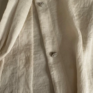 Beige blus med pärlknappar från Zara - Supersöt beige blus från Zara med volangkrage och knyt framtill. Blusen har långa ärmar med breda muddar och unika pärlknappar som ger en extra touch. Tillverkad i mjukt linne, perfekt för dig som gillar romantiska detaljer.