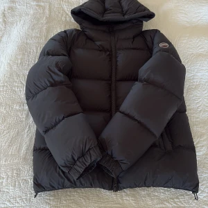 Svart pufferjacka från Colmar - Säljer nu min svart pufferjacka från Colmar med huva och markerad logga på ärmen. Jackan har en clean look, breda quiltade sektioner och dragkedja framtill. Perfekt för kalla dagar, med elastiska muddar och två sidofickor med dragkedja. Snygg och funktionell för vinter. Ny pris 6995