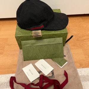 Svart Gucci keps med GG-mönster - Säljer en svart keps från Gucci i storlek M, tillverkad i Italien. Kepsen har det klassiska GG-monogrammet ton-i-ton och en snygg grön-röd rand på sidan. Justerbar rem bak i skinn och medföljer originalförpackning och certifikat. Perfekt för dig som gillar lyxiga accessoarer. Kvitto finns i papper och digitalt. Pris går att diskutera vid snabb affär