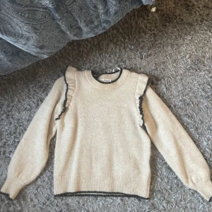 Beige stickad tröja med volang - Supersöt beige stickad tröja från Selected Femme med svarta volangdetaljer längs axlarna och kanten. Tröjan har rund hals och långa ärmar med ribbade muddar. Perfekt för dig som gillar en chill men ändå stylish look.