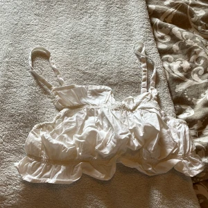 Vit volang linnetopp med smala axelband - Supersöt vit linnetopp med smala justerbara axelband och volangdetaljer. Toppen har en rynkad, croppad passform och är tillverkad i mjuk bomull. Perfekt för varma dagar och ger en chill vibe till din outfit.