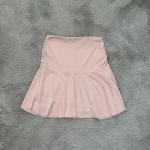 Ljusrosa/smutsrosa kortkjol med volang - Supersöt ljusrosa kortkjol med volangkant och rynkade detaljer på sidorna. Kjolen har en mjuk och flowy passform som ger en feminin vibe. Perfekt att styla med en crop top eller hoodie för en chill look.