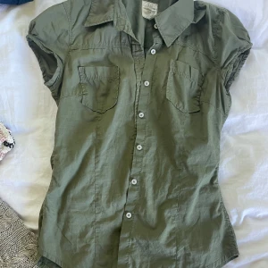 Olivgrön skjorta från L.O.G.G. H&M - Snygg olivgrön skjorta från L.O.G.G. H&M i storlek 34. Modellen har korta ärmar med lätt puff, två bröstfickor och klassisk krage..