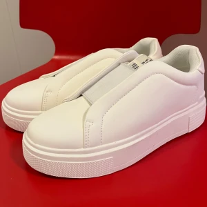 Vita sneakers - Säljer ett par stilrena vita sneakers med elastisk rem över vristen istället för klassisk snörning. Skorna har en chunky platt sula och är tillverkade i syntetmaterial med mjuk vadderad insida. Perfekt för dig som gillar minimalistisk och clean look. Endast testade men var tyvärr för stora för mig. Nypris 400kr säljer för 340kr