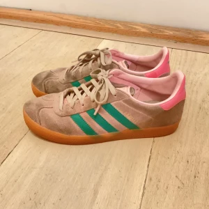 Adidas Gazelle sneakers rosa/grön - Adidas Gazelle sneakers i beige mocka med gröna ränder och detaljer i rosa på häl och plös. Klassisk låg modell med snörning och gummisula i brunt. Perfekt för dig som gillar retrovibbar och färgkombinationer som sticker ut.