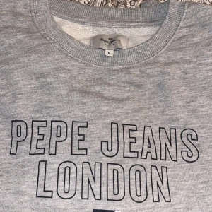 Grå sweatshirt från Pepe Jeans London - Snygg grå sweatshirt från Pepe Jeans London med stor svart logotyp på bröstet och en liten grafisk detalj i blått, vitt och rött. Rund hals och mjukt material som är perfekt för chill dagar. Passar dig som gillar enkel och stilren streetwear. Strl M i nyskick