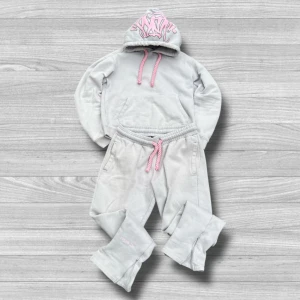 Syna World Tracksuit - Grå Syna World tracksuit Me med rosa snören och coolt rosa graffiti-tryck på huvan. Mjuk fleece på insidan och stor magficka framtill. Perfekt för chill dagar och streetwear-vibe. Snyggt broderad text på benet och matchande tryck på byxorna. Storlek L men passar M