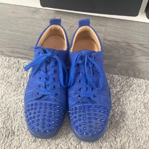 Blå Christian Louboutin sneakers med nitar - Säljer ett par blå sneakers från Christian Louboutin med coola nitar på tån och klassisk röd sula. Skorna har snörning och är tillverkade i mocka med en rund tå. Perfekt för dig som vill sticka ut med en unik och edgy stil. Har haft den sen våren 2024 men använder dom knappt längre.