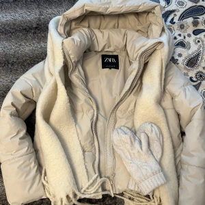 Beige pufferjacka från Zara med huva - Snygg beige pufferjacka från Zara med stor huva och två fickor med dragkedja. Jackan är quiltad och har en loose passform som ger en chill vibe. Perfekt för kalla dagar och enkel att matcha med din stil. Dragkedja framtill för smidig stängning.