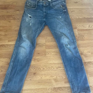 Blå Replay jeans med slitningar, stl 34 - Snygga blå jeans från Replay i storlek 34 med coola slitningar och distressed detaljer på benen. Klassisk femficksmodell med bruna läderpatch bak och tydliga Replay-sömmar på bakfickan. Jeansen har en rak passform och är tillverkade i slitstarkt denim.