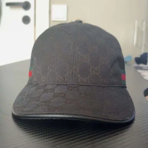 Svart Gucci keps med GG-mönster - Snygg svart keps från Gucci med diskret GG-monogram över hela kepsen och klassiska gröna och röda ränder på sidorna. Justerbar rem baktill för perfekt passform. Materialet är bomull och kepsen har en böjd skärm för en cool look., PRIS DISKUTERAS 
