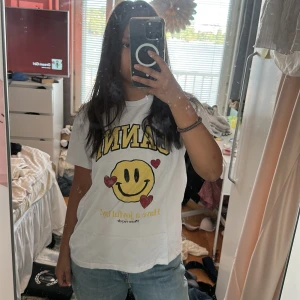Vit t-shirt från Ganni med smiley - Säljer en vit t-shirt från Ganni med stort tryck av en gul smiley och röda hjärtan på framsidan. T-shirten har korta ärmar och rund hals. Perfekt för dig som gillar coola prints och vill sticka ut lite extra.