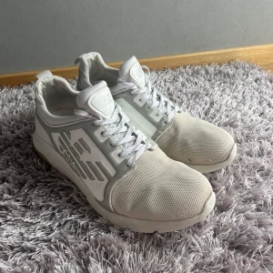 Vita EA7 Emporio Armani sneakers - Snygga vita sneakers från EA7 Emporio Armani med grå och vita detaljer. Skorna har snörning och en rund tå, samt en platt sula. Ovandelen är i mesh och syntetmaterial för en sportig vibe.