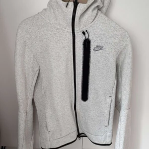 Ljusgrå Nike hoodie med dragkedja XS - Nike hoodie i ljusgrått med svart dragkedja och huva. Snyggt broderad Nike-logga på bröstet och en vertikal ficka med svart kant. Långärmad och tillverkad i mjukt bomullsmaterial, perfekt för chill eller träning. Passar dig som gillar sportig och clean stil.