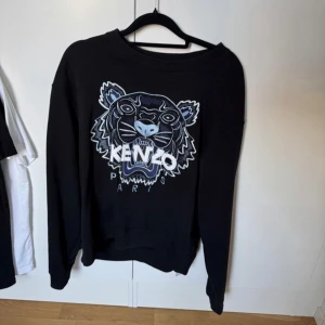 Svart Kenzo sweatshirt med tigertryck - Svart sweatshirt från Kenzo med stort broderat tigerhuvud och logga i vitt och blått på bröstet. Tröjan har rund hals, långa ärmar och ribbade muddar. Perfekt för dig som gillar statement-plagg och coola prints.