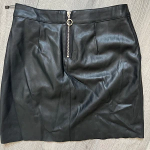 Svart skinnkjol med dragkedja - Snygg svart kort skinnkjol med en cool silverfärgad dragkedja framtill och stilrena sömmar. Kjolen har en rak passform och är tillverkad i mjukt skinnmaterial som ger en edgy vibe. Perfekt för dig som gillar minimalistisk stil med en twist.