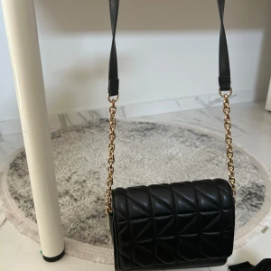 Svart quiltad axelväska från MNG - Snygg svart axelväska från MNG med quiltad framsida och guldfärgad kedja som axelrem. Väskan har en stilren, rektangulär form och är tillverkad i skinnimitation. Perfekt storlek för det viktigaste och ger en cool vibe till din outfit.