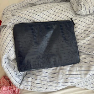 Mörkblå kuvertväska i skinn - Snygg mörkblå kuvertväska med croco-mönstrad yta och guldfärgad dragkedja. Perfekt storlek för laptop eller dokument, stilren och enkel att matcha med olika outfits. Materialet är skinn och väskan har ett minimalistiskt, rektangulärt format.
