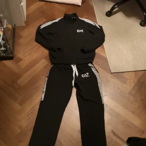 EA7 tracksuit  - Snyggt svart träningsset från Emporio Armani EA7 med vita detaljer och logga på både jacka och byxor. Jackan har dragkedja och ståkrage, byxorna har snörning i midjan och vita ränder längs sidorna. Perfekt för dig som vill ha stil även på gymmet.