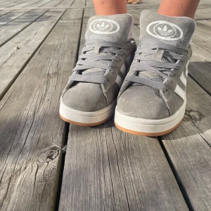 Supersöta gråa campus💗 - Säljer ett par grå Adidas Campus sneakers med klassiska vita ränder på sidan och Adidas-logga på plösen. Säljs då dem inte längre passar💗 Vid hälarna är det en slitning!!!💗 Kom gärna med prisförslag. Köpta för 1500 kr! 