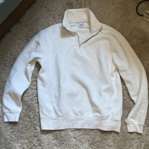 Vit half zip tröja från Zara - Säljer en stilren vit tröja från Zara med halv dragkedja och hög krage. Tröjan har långärmade ärmar och är gjord i mjuk bomull, perfekt för chill dagar. Enkel design utan mönster och med ribbade muddar vid ärmslut och nederkant.