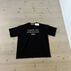 Svart Acne Studios t-shirt Stockholm M - Svart t-shirt från Acne Studios med vit text 'Acne Studios STOCKHOLM 1996' på bröstet. Klassisk rund hals och korta ärmar. Materialet är mjuk bomull och passformen är loose, perfekt för en chill och stilren look. Kommer med tag.