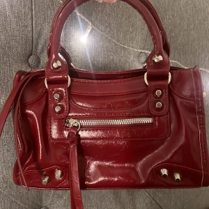Vinröd handväska med silverdetaljer - Säljer en vinröd handväska, man kan även ha den som axelbandsväska. Väskan har både handtag och axelrem, så du kan bära den på flera sätt. Det följer med längre axelband också. Mått: 15x8x24cm. Aldrig använd och är i helt nytt skick.