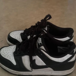 Nike Dunk Low svartvita sneakers - Svartvita Nike Dunk Low sneakers med klassisk siluett och perforerad tåbox. Skorna har svart swoosh, svarta snören och vit bas. Låg modell med platt sula. Skorna har små skador på innersidan och där framme. Kolla bilderna!