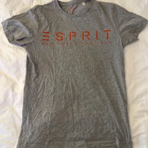 Grå t-shirt från Esprit med tryck - Snygg grå t-shirt från Esprit med rött tryck över bröstet där det står 'ESPRIT San Francisco 1968'. Klassisk rund hals och korta ärmar, perfekt för chill dagar. Mjuk och skön bomull som känns nice mot huden.