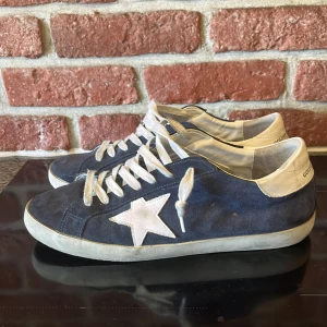 Mörkblå Golden Goose sneakers mocka - Snygga mörkblå sneakers från Golden Goose med vit stjärndetalj på sidan och GGDB/SSTAR-tryck i guld. Skorna har vit platt sula, vita skosnören och detaljer i vitt skinn vid häl och tå. Tillverkade i mocka för en cool och avslappnad look. Hör av er vid frågor eller prisförslag