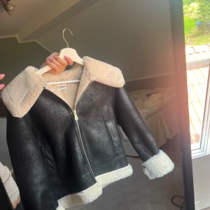 Svart teddyjacka från Mango - Snygg svart teddyjacka från Mango med krage, muddar och insida i beige fuskpäls. Jackan har dragkedja framtill och två fickor. Perfekt för kalla dagar när du vill vara både varm och stilren. Ny pris 1200kr