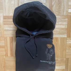Svart Polo Bear hoodie från Ralph Lauren - Svart hoodie från Polo Ralph Lauren med broderad Polo Bear på bröstet. Klassisk huva med snörning och mjukt material. Perfekt för dig som gillar streetstyle och vill ha något ikoniskt i garderoben.