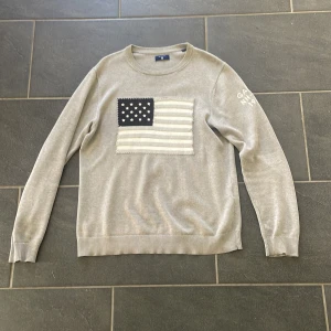 Grå stickad tröja med flagga från GANT - Snygg grå stickad tröja från GANT i storlek M. Framtill finns en broderad amerikansk flagga i vitt och mörkblått, och på ena ärmen står det 'GANT NH-1949' broderat. Tröjan har rund hals och långa ärmar med ribbade muddar.