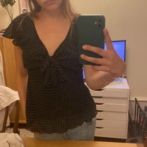 Svart prickig blus från H&M - Supersöt svart blus från H&M med små vita prickar, v-ringning och knyt framtill. Blusen har korta, volangiga ärmar och vågig nederkant. Perfekt att styla med jeans eller kjol!