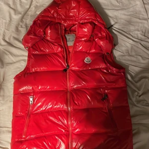 Röd dunväst från Moncler med huva - Säljer en riktigt snygg röd dunväst från Moncler, Västen har två fickor med dragkedja, Moncler-logga på bröstet och quiltad design. Perfekt för en höst dag eller en sommarkväll. Osäker om den är äkta för varje gång jag försöker skanna på moncler appen så låter den mig inte logga in