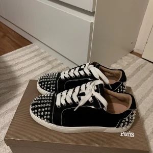 Christian Louboutin svarta sneakers med nitar - Svarta sneakers från Christian Louboutin med vita snören och ikonisk röd sula. Skorna har silvriga nitar på tå och häl för en edgy vibe. Ovandelen är i mocka och detaljer i skinn, perfekt för dig som vill sticka ut. 