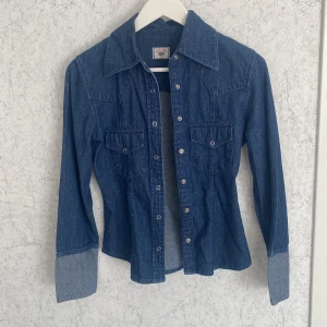 Jeansskjorta med tryckknappar - Snygg jeansskjorta från Gold Label i klassisk blå denim. Skjortan har markerad krage, långa ärmar med breda manschetter och tryckknappar framtill. Två bröstfickor med lock och en figurnära passform ger en cool och tidlös vintage look. Storlek 36.