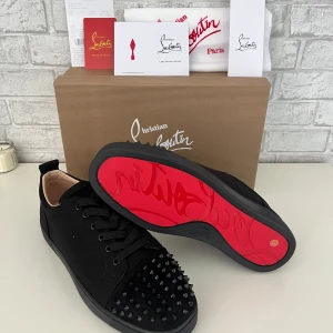 Christian Louboutin svarta sneakers med nitar - Svarta sneakers från Christian Louboutin med mattsvarta nitar på tån och ikonisk röd sula. Skorna har snörning, rund tå och är tillverkade i skinn och mocka. Perfekt för dig som vill sticka ut med en lyxig och edgy stil.