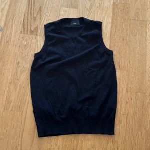 Svart stickad väst från Next - Klassisk svart stickad väst från Next med v-ringad hals och ribbade kanter. Perfekt att slänga över en skjorta för en clean och stilren look. Enkel design utan mönster, passar till det mesta och ger en snygg layering-effekt.