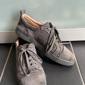 Grå sneakers från Christian Louboutin - Säljer ett par grå sneakers från Christian Louboutin med klassisk röd sula och signatur på undersidan. Skorna har snörning, rund tå och är tillverkade i mocka med beige skinnfoder. Metallöglor i guld och silver ger en cool kontrast. Perfekt för dig som gillar exklusiva street vibes.