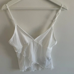Vit mesh topp - Supersöt vit mesh topp  med tunna axelband och v-ringad front. Toppen har dubbla lager mesh med volangdetaljer som ger en drömmig och luftig vibe. Perfekt för dig som gillar romantiska och drömlika plagg.