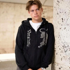 Svart Von Studio hoodie med tigertryck - Svart hoodie från Von Studio med glittrande tigertryck och logga på framsidan. Tröjan har huva men snöret runt är borta, dragkedja och två fickor framtill. Materialet är mjuk bomull och passformen är normal. Perfekt för dig som gillar streetwear med lite edge.