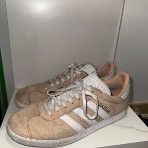 Beige/rosa Adidas Gazelle sneakers - Säljer ett Adidas Gazelle sneakers i ljus rosa mocka med vita detaljer. Knappast använda, kan rengöras vid köp