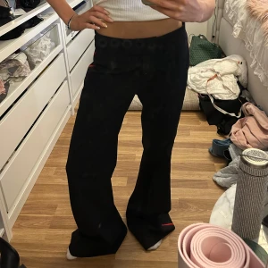 Svarta kostymbyxor Hunkydory med röd rand - Svarta kostymbyxor från Hunkydory med raka, vida ben och en röd rand längs sidorna. Byxorna ha diskreta fickor och stängs med dragkedja och hyska. Snygg detalj med uppvikta benslut. Perfekta för en trendig och cool look.