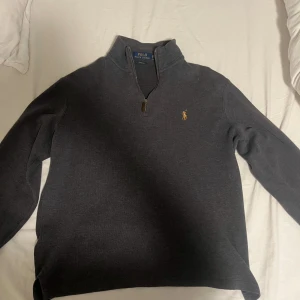 Mörkgrå half zip tröja Polo Ralph Lauren - Mörkgrå långärmad tröja från Polo Ralph Lauren i storlek XS. Klassisk half zip med ståkrage och broderad logga på bröstet. Tillverkad i mjuk bomullsmix, perfekt för lager-på-lager och chill stil.