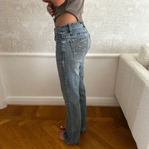 Lågmidjade blå jeans med broderi - Snygga lågmidjade jeans i ljusblå tvätt med raka ben och broderade bakfickor. Jeansen har klassisk femficksdesign, kontrastsömmar och coola detaljer på fickorna. Perfekta för dig som gillar en avslappnad och trendig look.