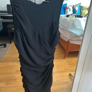 Svart långklänning från Mango Suit - Snygg svart långklänning från Mango Suit i storlek M. Klänningen har draperade detaljer framtill och baktill, en markerad midja och en synlig dragkedja i ryggen. Materialet är mjukt och följsamt, perfekt för dig som vill ha en elegant och stilren look.