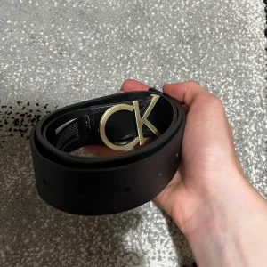 Svart Calvin Klein skärp med guldfärgat spänne - Snyggt svart skärp från Calvin Klein i äkta skinn med ett stilrent guldfärgat CK-spänne framtill. Bältet har en clean look med präglad logga på remmen och passar perfekt till jeans eller kostymbyxor. Längd 90 cm.